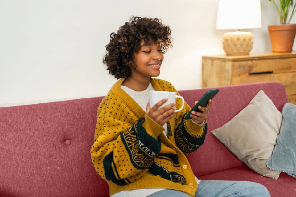 woman on couch checking phone and holding coffee